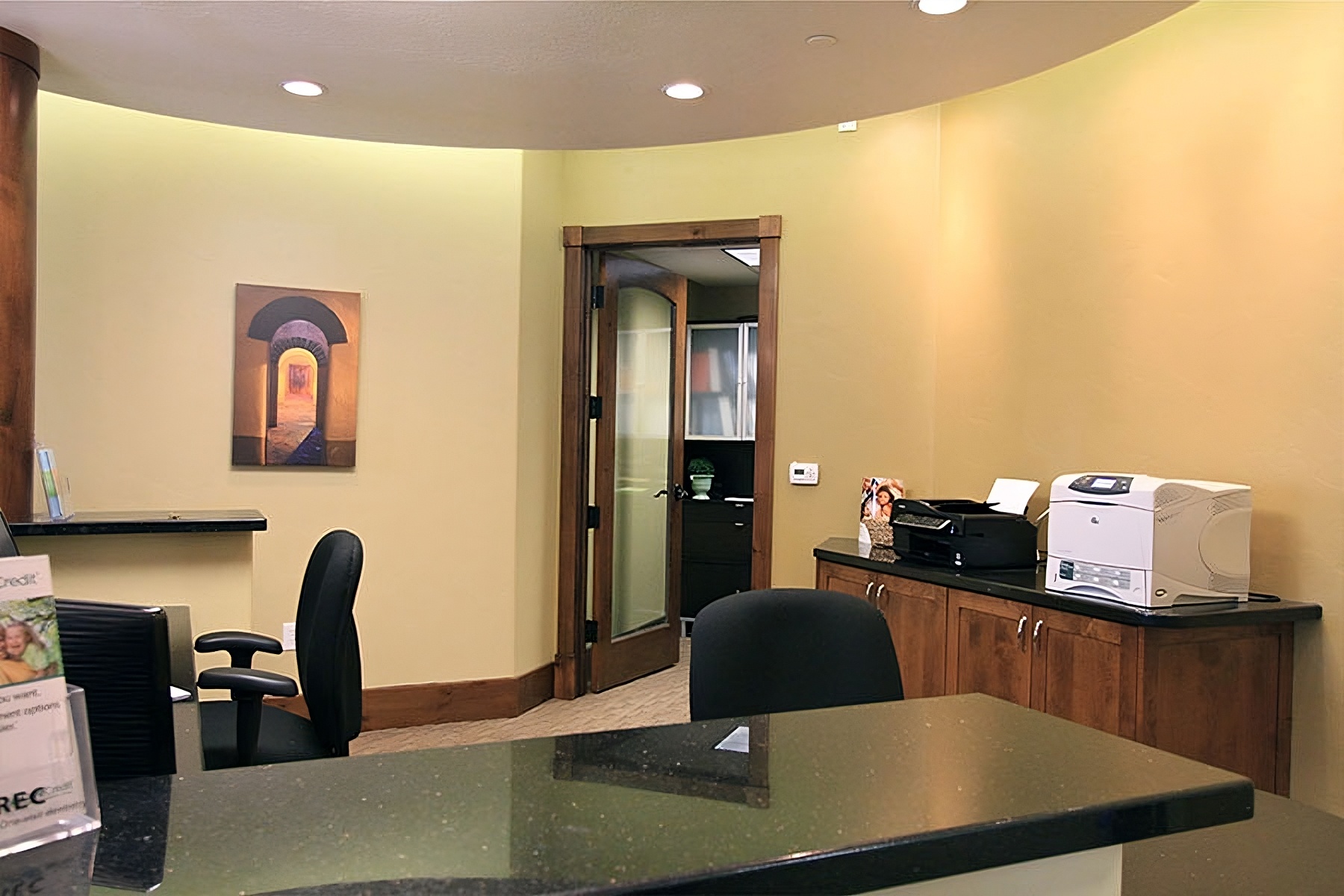 The image shows an interior view of an office reception area with a modern design, featuring a curved wall, a desk with a chair, a computer monitor, and a printer.