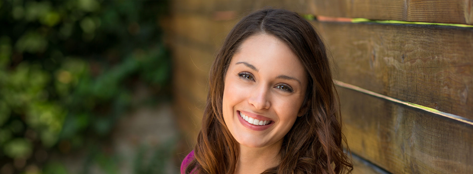A smiling woman posing in front of a wooden fence with greenery behind her.