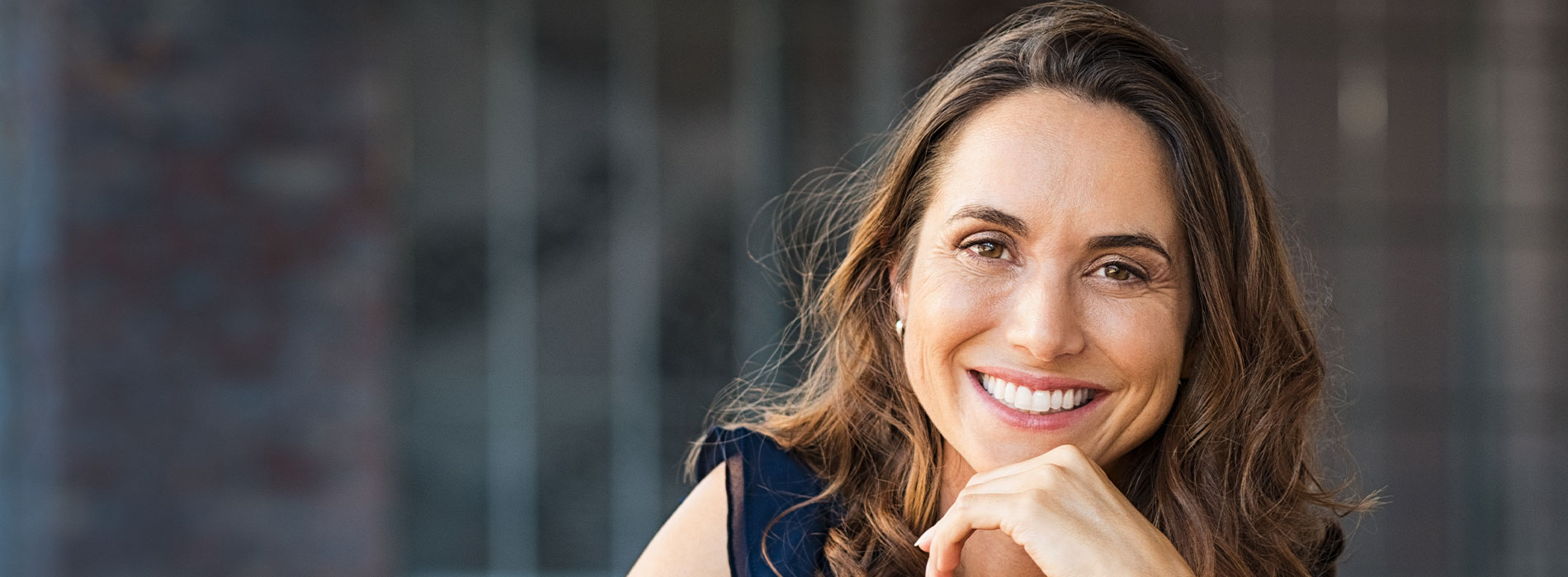 The image shows a woman with a smile, looking directly at the camera, against a blurred background.