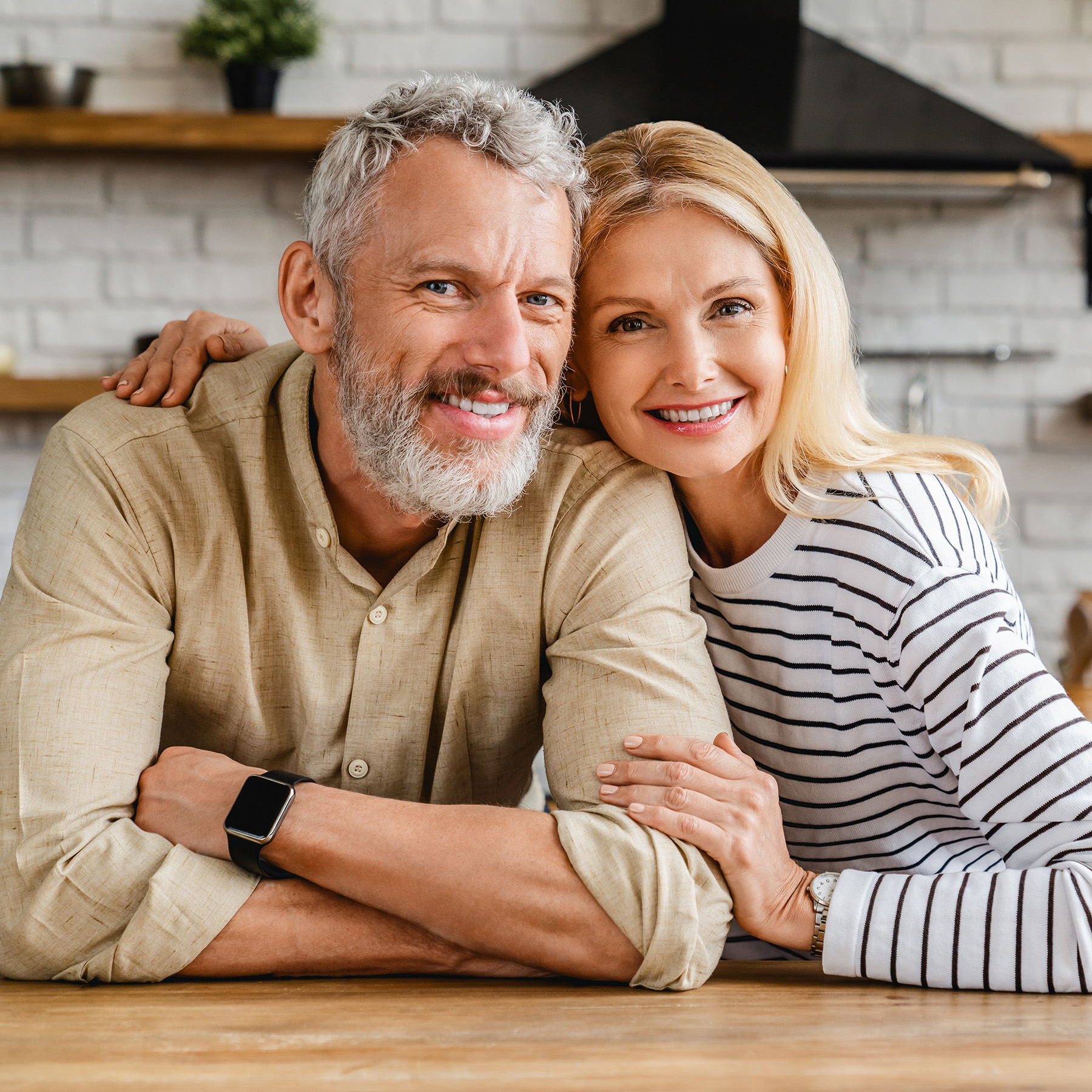 The image shows a man and woman posing closely together, smiling at the camera, with the man wearing a beard and the woman with blonde hair, both dressed casually, standing next to each other against a backdrop of a kitchen setting with a warm ambiance.