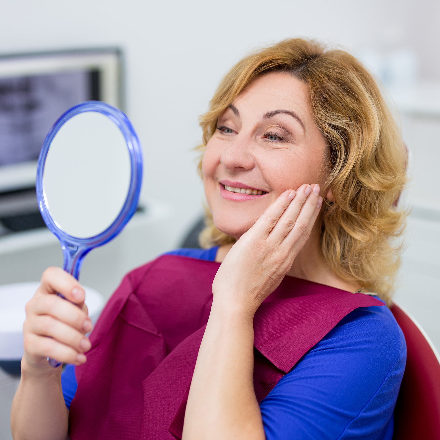 A woman with blonde hair, wearing a blue face mask, is seated in a dental chair, holding a mirror up to her face while looking directly at the camera.