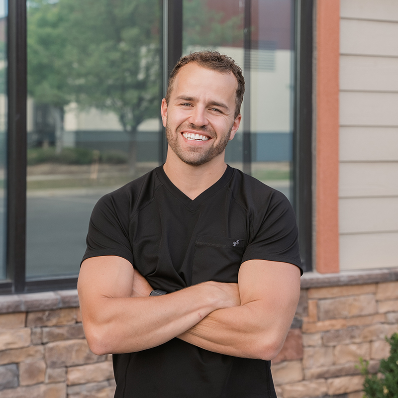 The image shows a man standing confidently with his arms crossed, wearing a black t-shirt and smiling at the camera. He appears to be indoors, possibly in a professional setting, given the background that includes a brick wall and a window with blinds partially drawn.