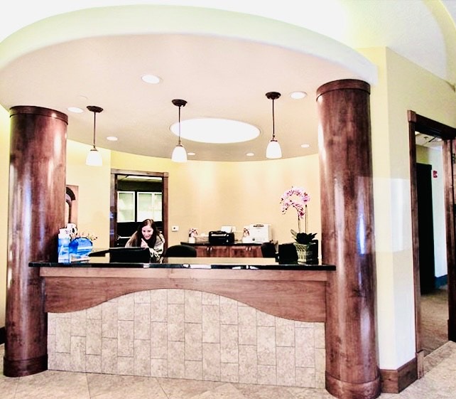 The image shows a well-lit interior space with a reception desk area featuring a countertop with a computer monitor, a person seated behind the counter, and a sign that reads RECEPTION . The room has a curved ceiling with recessed lighting and wood columns framing the space. The design is modern with earthy tones and a warm ambiance.