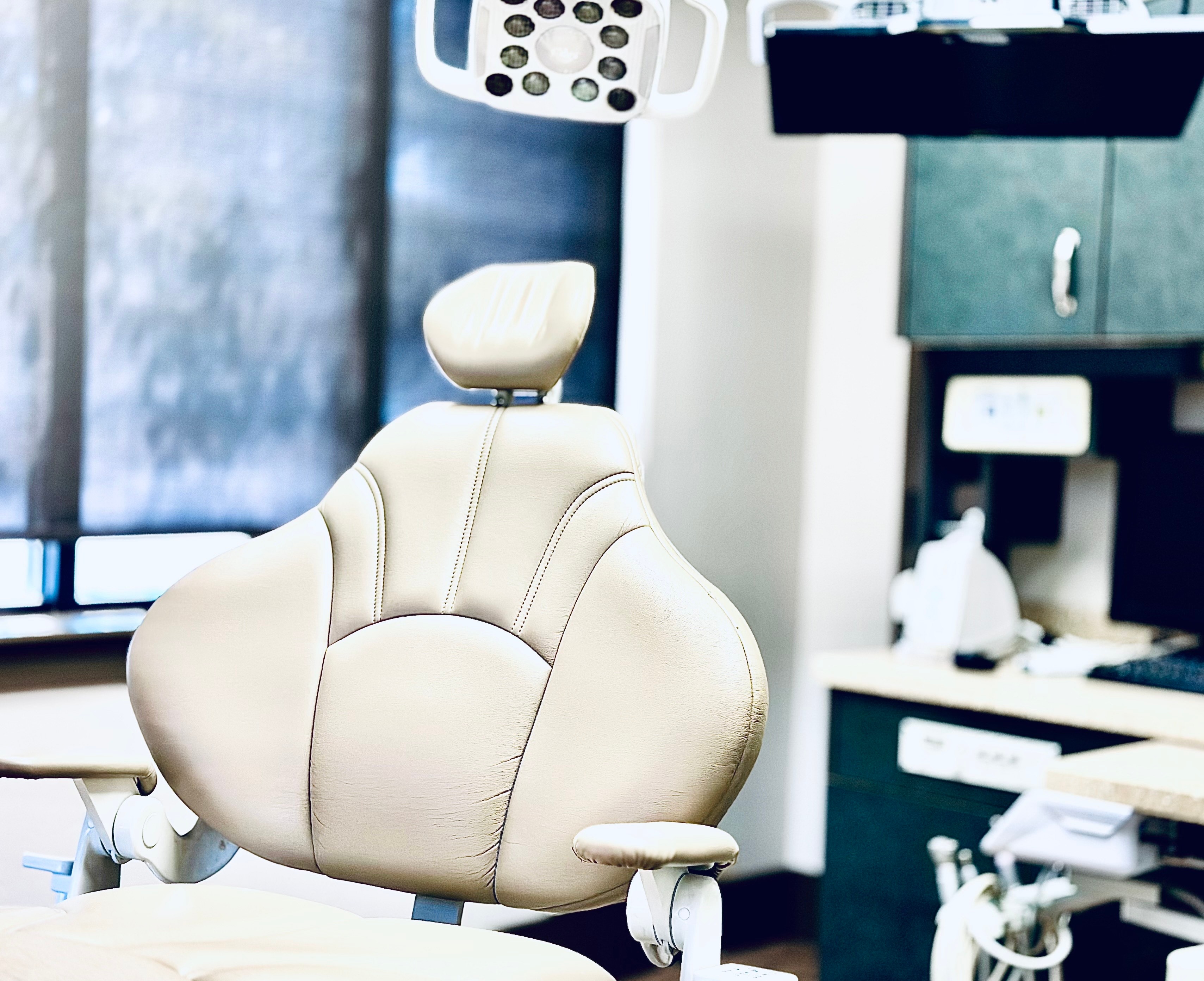 The image shows a dental chair inside a dental office, with medical equipment and a desk with a computer monitor visible in the background.