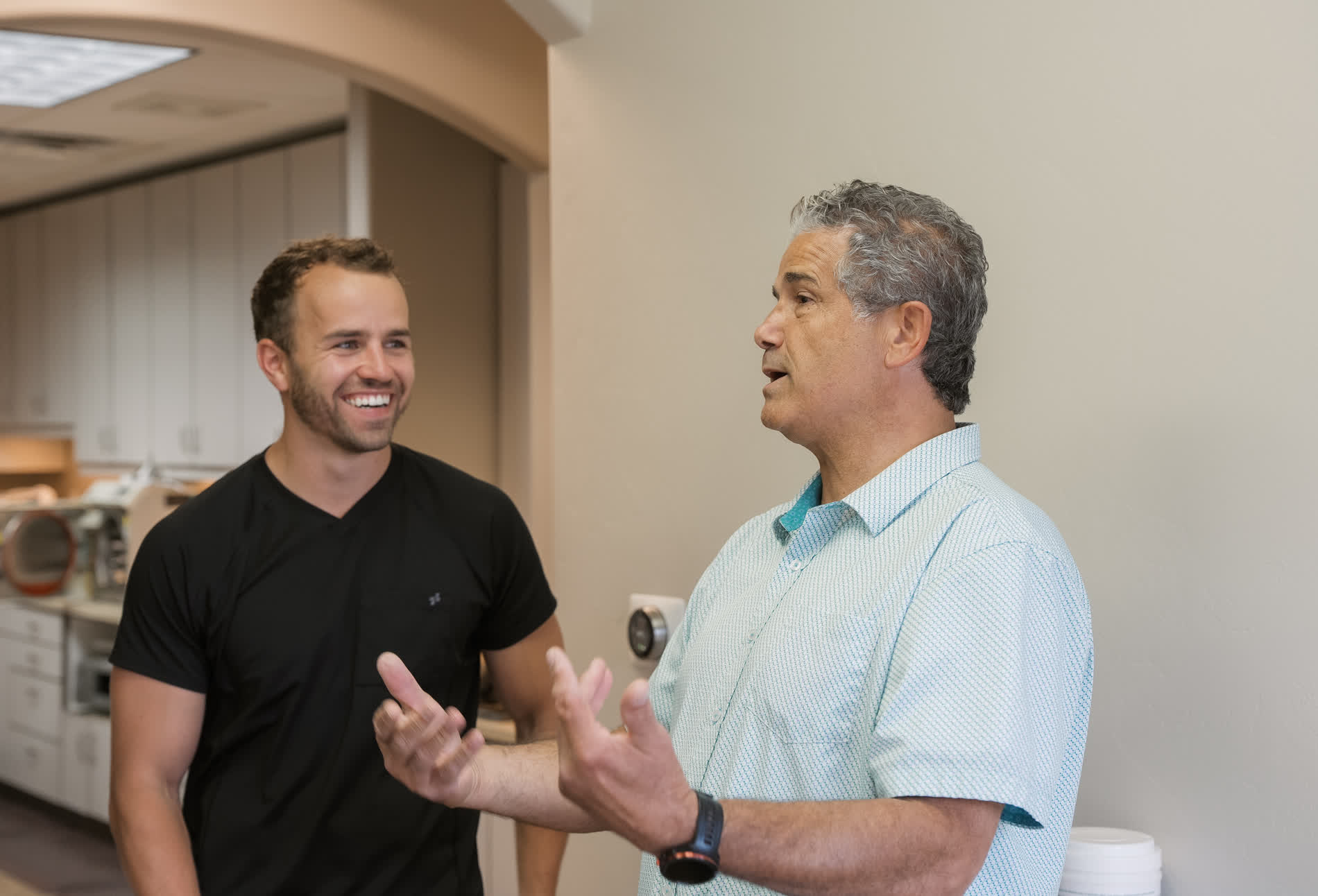 In the image, there are two men standing in an office environment. The man on the left appears to be gesturing with his hands while talking to the other man who is listening attentively.