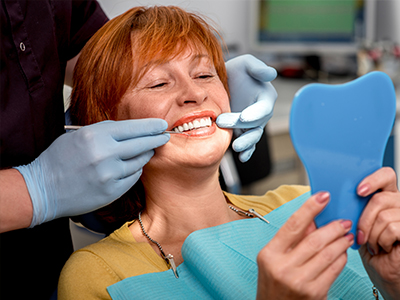 A woman with red hair, wearing glasses, sitting in a dental chair, holding up a blue toothbrush-shaped object, smiling at the camera while undergoing dental treatment.