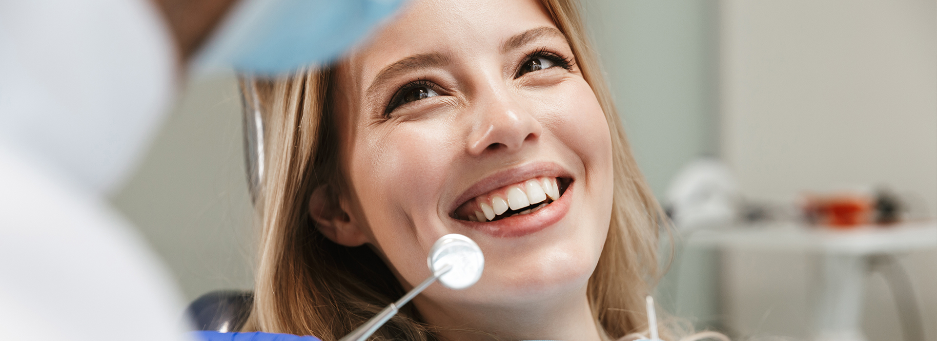 A woman with a radiant smile is seated at a dental chair, while a dentist in the background checks her teeth with a magnifying glass.