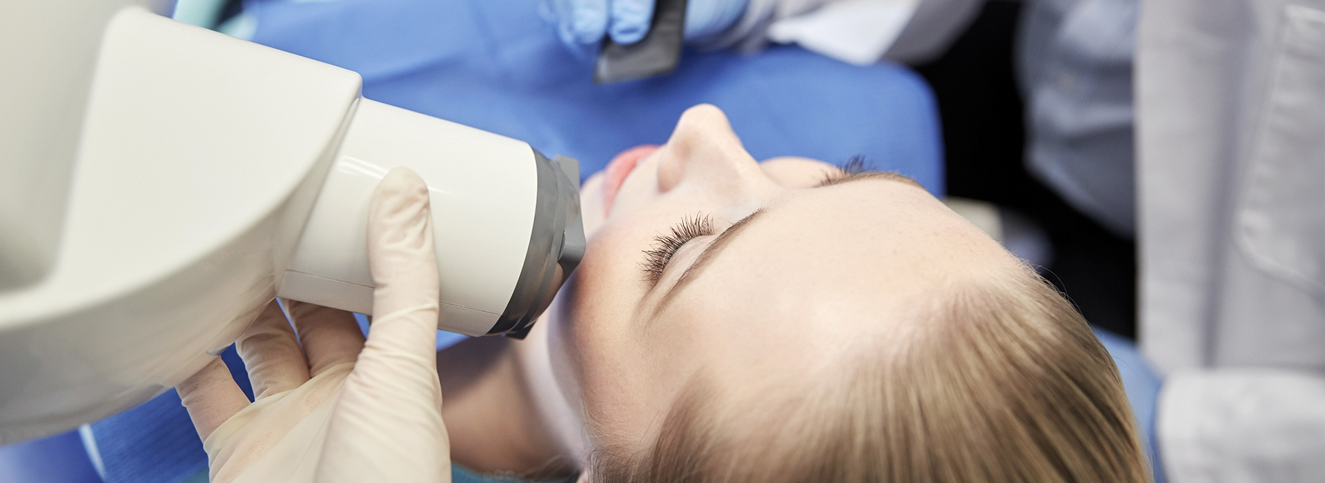This image shows a person receiving a medical procedure with a device attached to their face, while another individual, possibly a healthcare professional, appears to be operating the equipment, set against a blue backdrop that suggests a clinical environment.