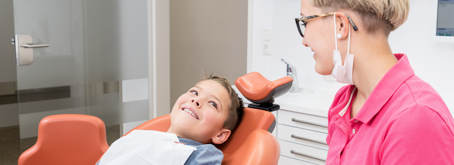 The image shows three people in a dental office setting a young boy sitting in a dental chair with an open mouth, a woman standing nearby wearing glasses and a pink shirt, and another person partially visible on the right side of the frame, all against a backdrop that includes dental equipment and a window.