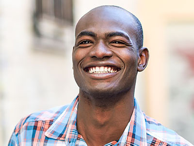 The image shows a smiling man with short hair, wearing a plaid shirt, standing outdoors in front of a building with shutters.