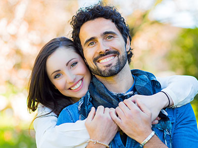 A man and woman embrace each other with smiles, standing outdoors during daylight.
