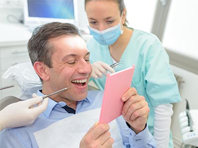 The image shows a man sitting in a dental chair with a pink cardboard sign on his face, smiling at the camera, while a dentist and a dental hygienist work around him.