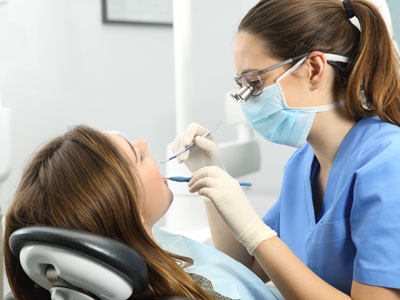 The image shows a dental hygienist performing a cleaning procedure on a patient s teeth while wearing personal protective equipment, including gloves and a face mask.