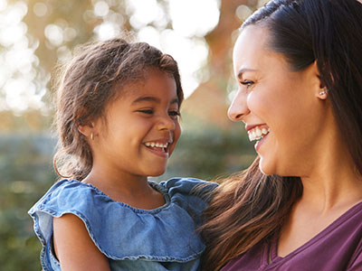 The image shows a woman standing outdoors with a young child, both smiling and looking towards each other.