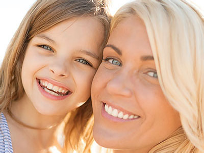 The image shows a woman with her child, both smiling at the camera, set against a blurred background which suggests an outdoor setting, possibly during daylight hours.