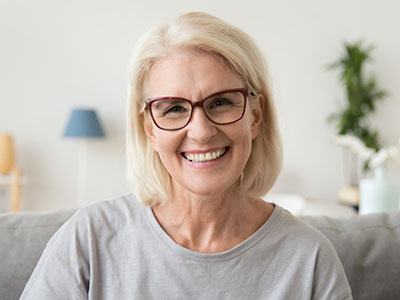 The image features a middle-aged woman with blonde hair, wearing glasses and a light-colored top, smiling at the camera. She has short hair and appears to be indoors, possibly in a living room setting, given the presence of a couch and a lamp in the background.