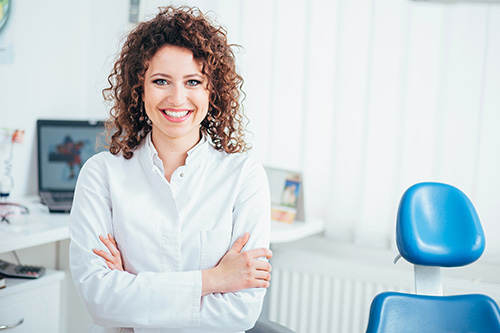 A woman with curly hair stands confidently in an office setting, leaning on her hands with a smile, wearing a white shirt.