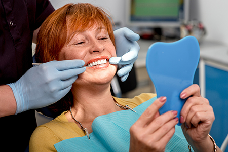 Woman sitting in dental chair with blue mouthguard, smiling at camera while holding it up with both hands.
