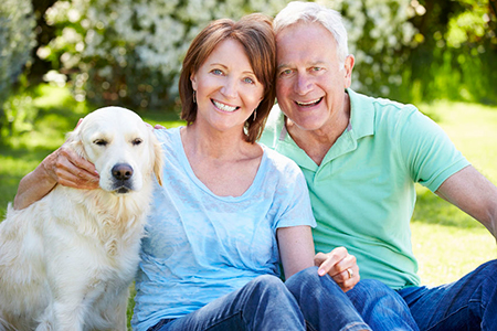 The image depicts an older man and woman sitting on grass with a golden retriever dog between them, smiling at the camera.