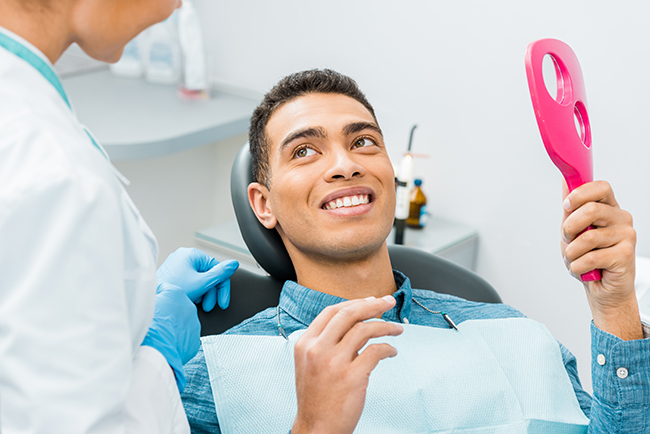 A man seated in a dental chair, smiling at the camera with a pink object in his hand.