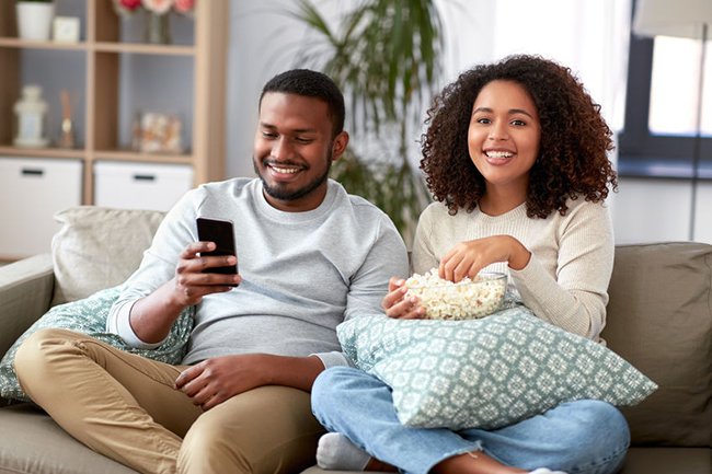 A man and woman are sitting on a couch, enjoying each other s company while watching television, with snacks visible nearby.