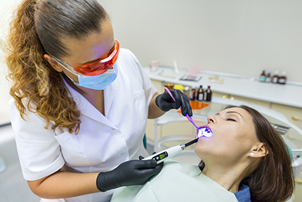 A dental hygienist is performing a procedure on a patient s teeth using a dental drill and a blue light, with the patient wearing a surgical mask and lying in a dental chair.