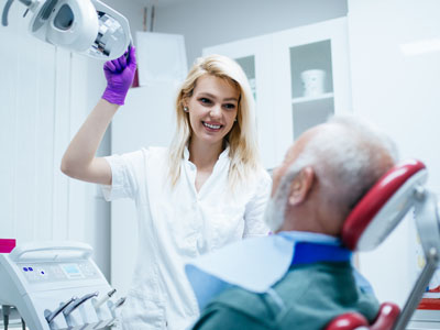A woman in a white coat stands next to an elderly man with a medical device on his face, who appears to be receiving dental care.