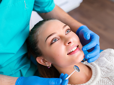 A woman receiving dental care from a professional, with her eyes closed and smiling, while being attended to by a dental hygienist wearing gloves and a mask.