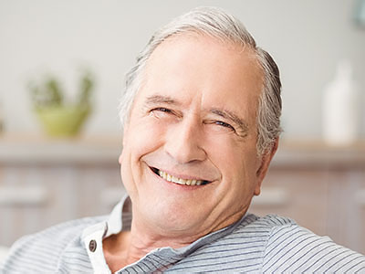 The image shows a smiling older man with gray hair, wearing a dark shirt, sitting comfortably with his head resting on one hand.
