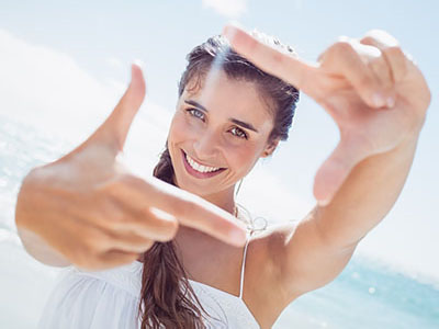 A woman with long hair is holding her hand up to frame a view, smiling at the camera while standing on a beach with clear skies.